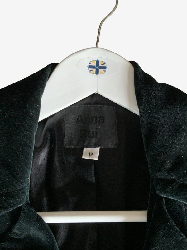 A close-up of a black velvet jacket hanging on a white hanger against a light gray background. The inner label reads 'Anna Sui' and features a small blue and yellow cross logo on the hanger.