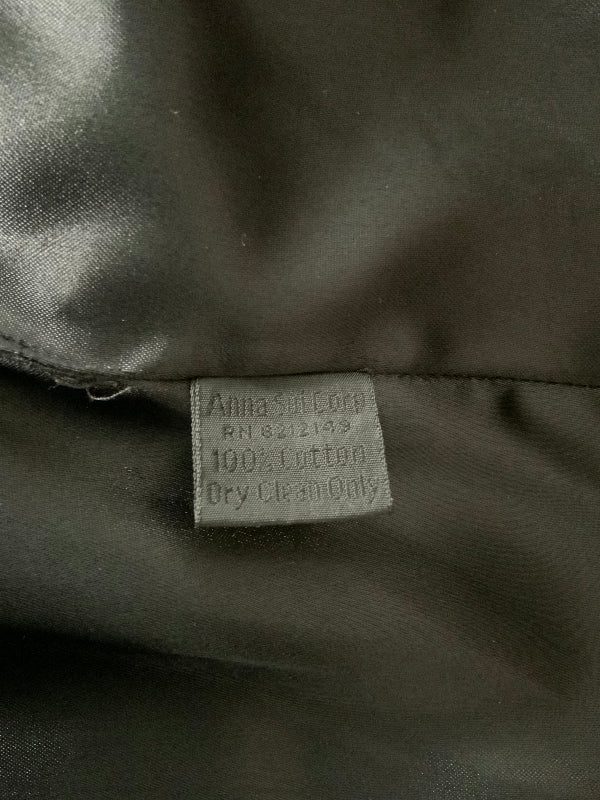 Close-up of a black or black fabric tag sewn onto a shiny dark satin-like surface. The tag reads 'Anna Sui Cotton' and '100% Cotton Dry Clean Only'.