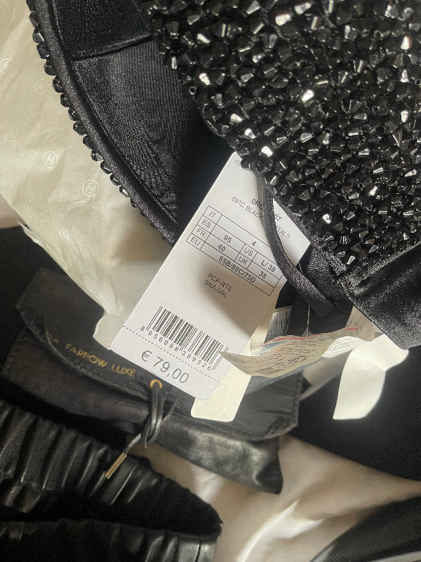A close-up of a black shoe with a heavily embellished black beaded upper, attached to a white size and price tag, resting on white tissue paper next to a black shoe bag.