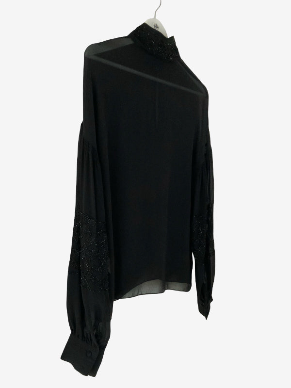 A black long-sleeve blouse with sheer mesh panels and textured beaded trim on the collar and cuffs, hanging on a white hanger against a light gray background.