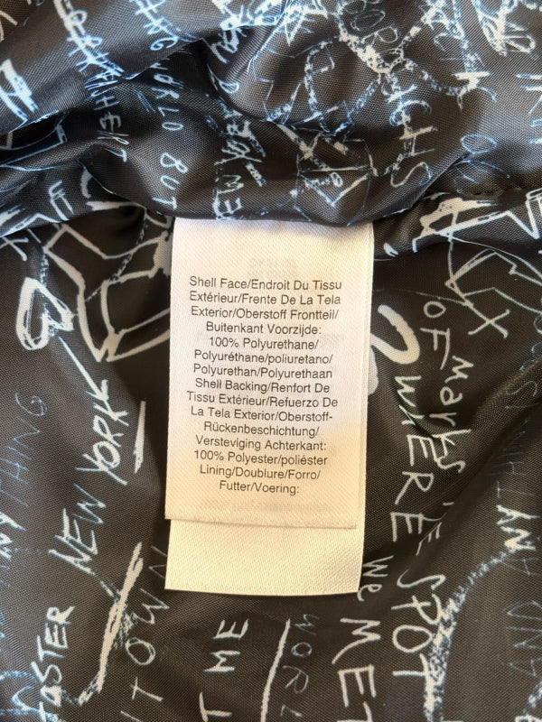 Close-up of a garment's interior label and lining. The lining is black with a white graffiti-style script print, and the central sewn-on paper label reads 'Shell Face/Endroit Du Tissu / Polyurethane / 100% Polyurethane'.