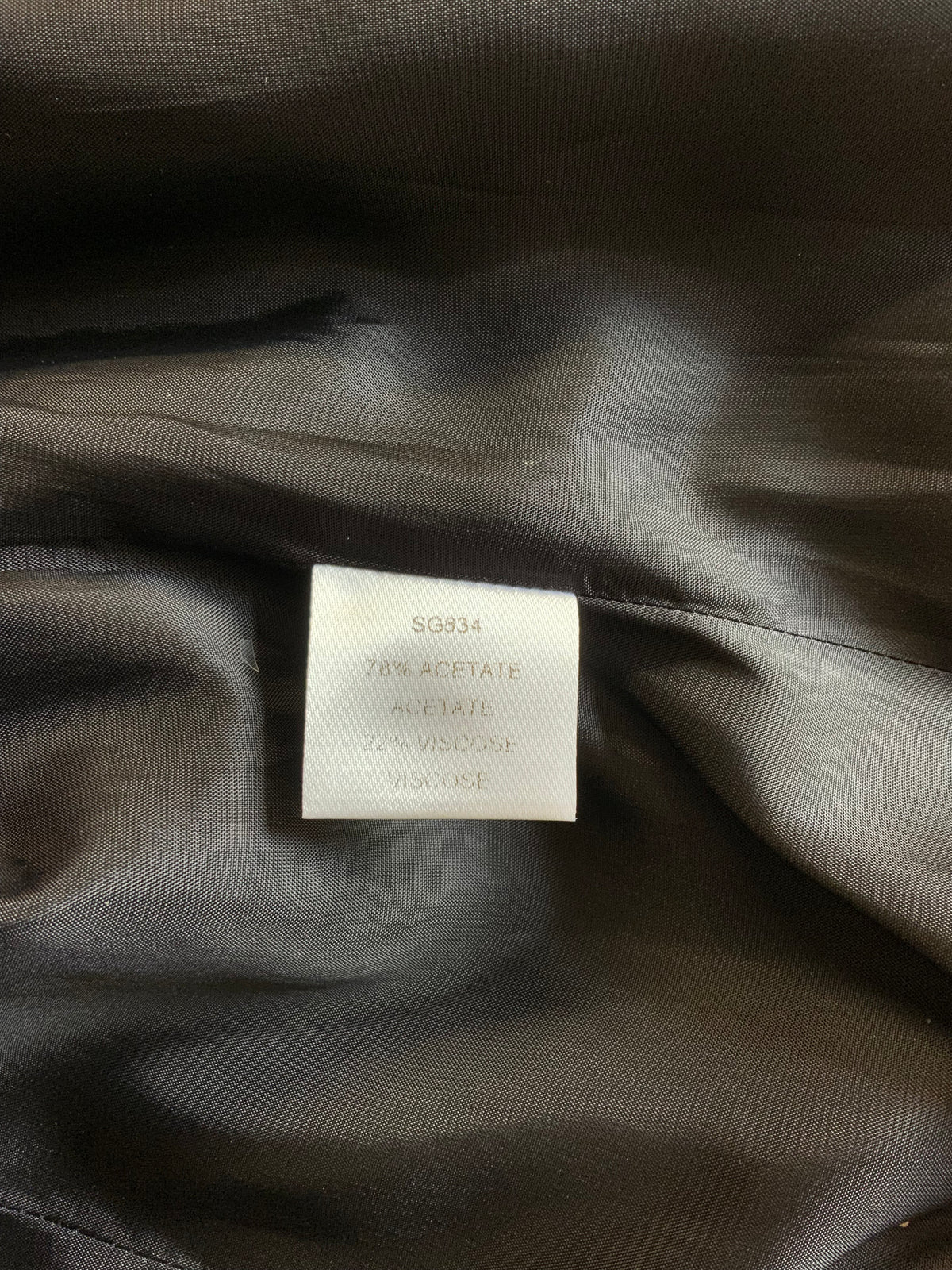 Close-up of a dark satin or silk-like garment interior with a small white size/composition label sewn into the fabric. The label reads SG834 78% ACETATE 22% VISCOSE