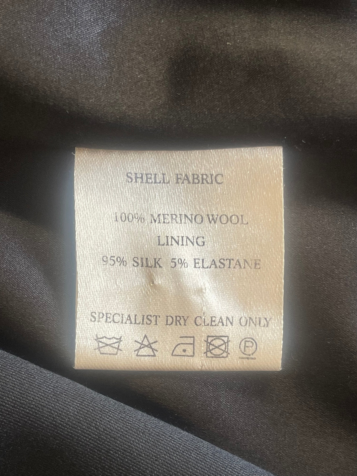 A close-up of a beige fabric care label inside a black garment, showing text that reads 'SHELL FABRIC 100% MERINO WOOL LINING 95% SILK 5% ELASTANE' with care symbols below.