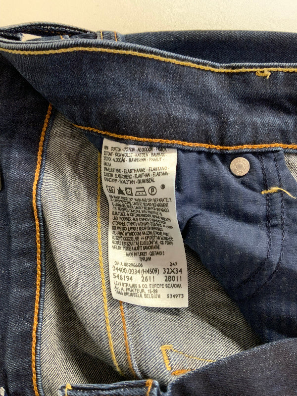 Close-up of the inside waistband of dark blue denim jeans, showing a white size and care label sewn onto the back of the waistband, with yellow contrast stitching and a metal button.
