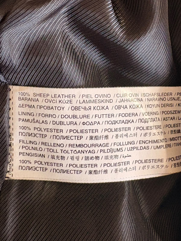 Close-up of a sewn-in product label on dark gray or black textured fabric, showing material composition text in multiple languages including sheep leather, polyester, and polyurethane.