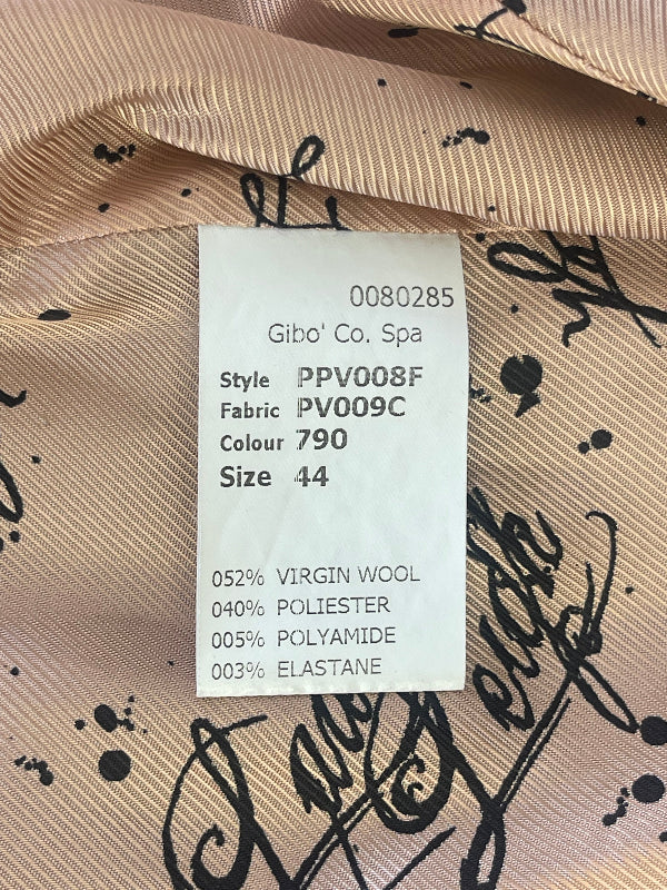 A close-up of a beige silk-like garment fabric with black script and splatter print, featuring a white paper label that reads 'Gibo' Co. Spa' and includes color and material details.