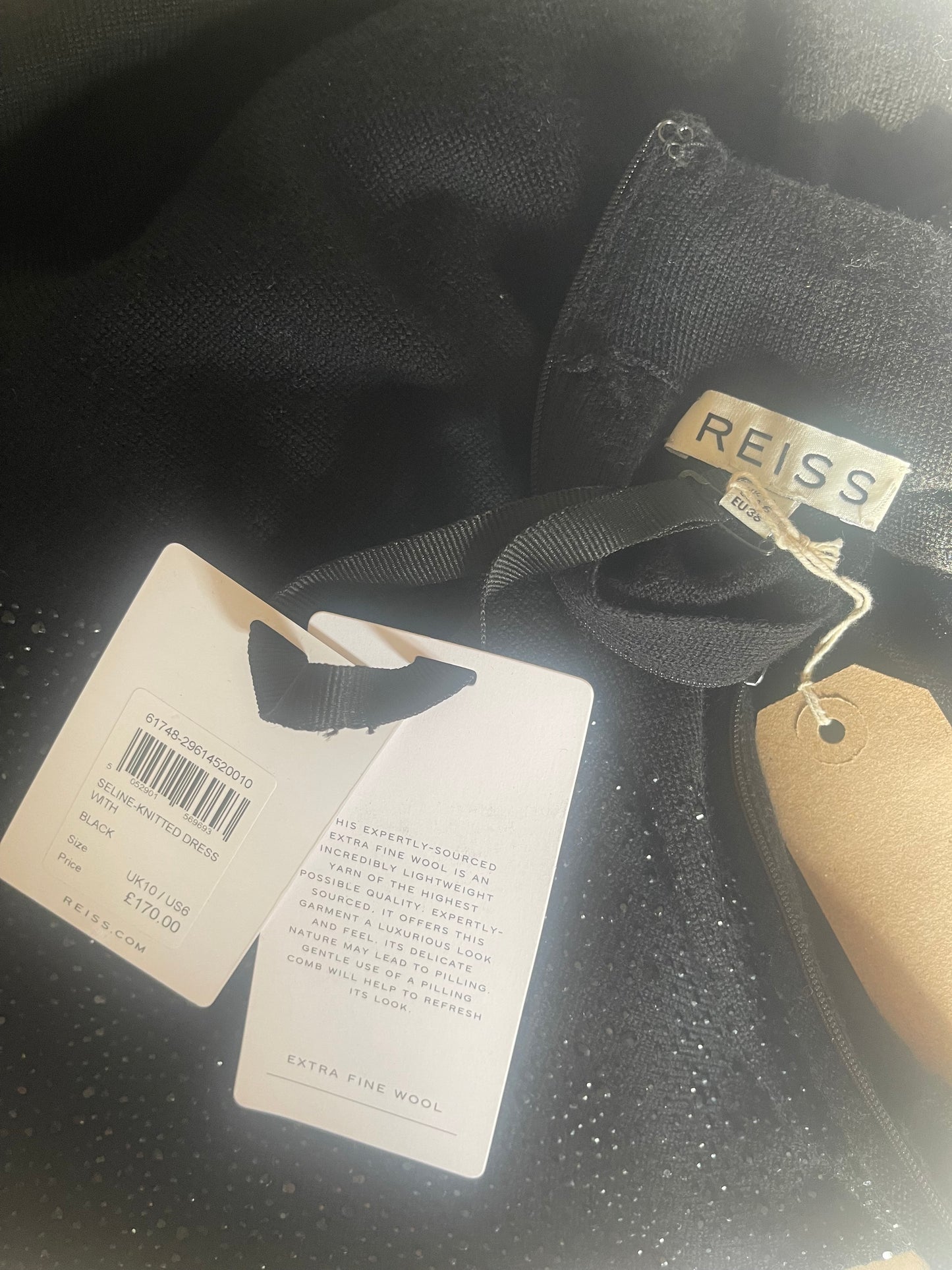 Close-up of a black garment with a visible REISS brand label, attached black ribbon, and white product tags on a dark textured surface.