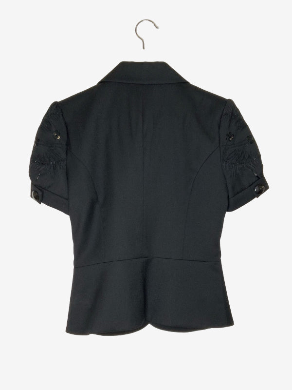 Black short-sleeve blazer shown from the back, hanging on a metal hanger against a light gray background. The blazer features structured shoulders with decorative embroidered details and small gold-tone button accents.