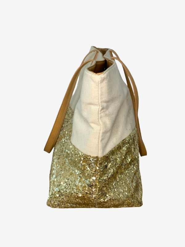 A color-blocked tote bag with a cream canvas upper section, a gold sequined lower section, and tan shoulder straps, shown against a plain light gray background.
