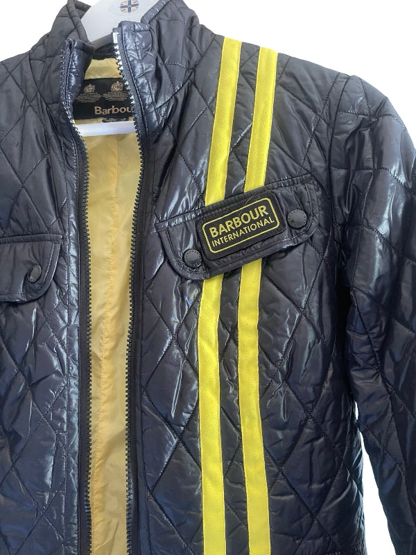 A close-up of a quilted Barbour jacket in black with two vertical yellow stripes and a yellow zipper, featuring a Barbour patch on the chest pocket.
