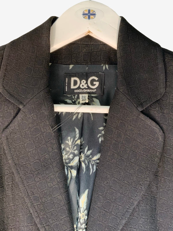 A close-up of a black textured silk blend blazer by Dolce & Gabbana, hanging on a white hanger with a floral-patterned silk lining. The D&G brand label is clearly visible inside the collar