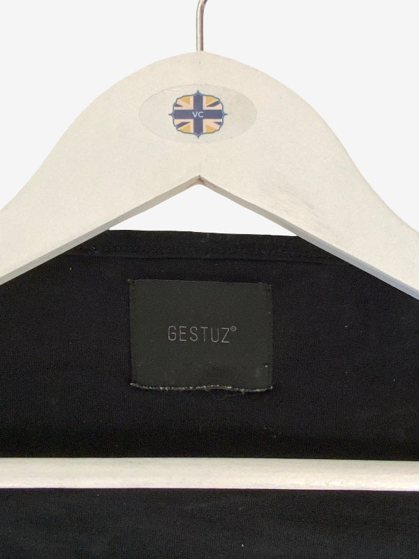 Close-up of a black garment hanging on a white wooden hanger against a white background. The hanger has a small circular logo near the top, and the garment label reads GESTUZ.