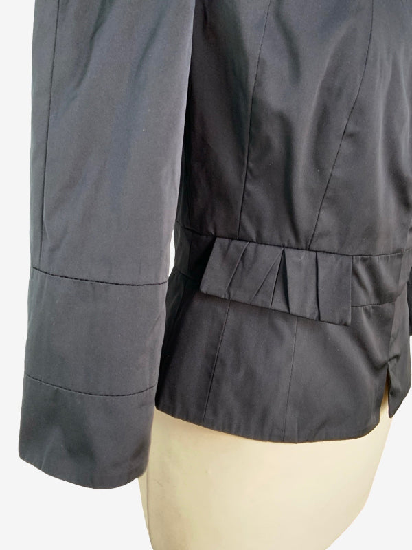 Close-up of a black stretch satin evening jacket displayed on a cream-colored mannequin torso, showing the sleeve cuff, pleated waist detail, and fabric texture.