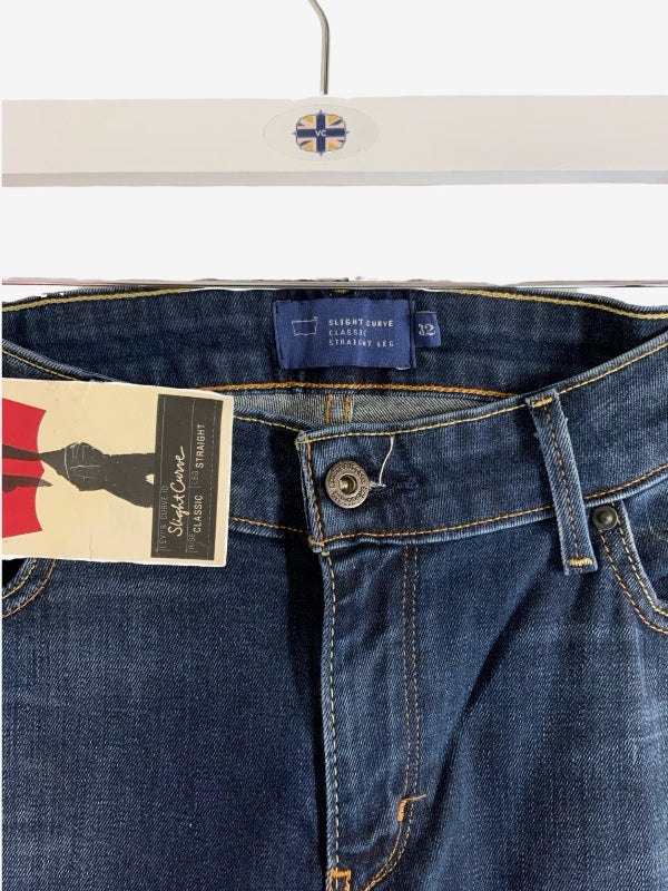 Close-up of a pair of dark blue denim jeans hanging on a white hanger, showing the waistband, button closure, belt loops, and a paper brand tag attached to the left side.