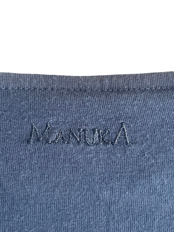 Close-up of a blue-gray ribbed knit fabric with the word 'MANUKA' embroidered across the chest, shown against a white background.