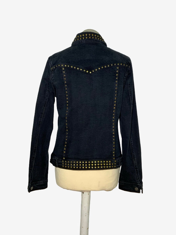 A dark wash denim jacket with gold-tone metal studs, shown from the back on a cream-colored dress form mannequin against a plain white background.