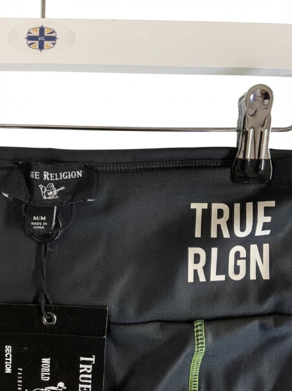 Close-up of black True Religion leggings clipped on a hanger, with large white 'TRUE RLGN' text on the front and a black hangtag attached.