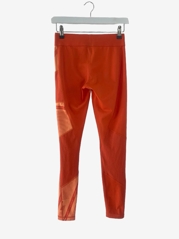 Bright orange athletic leggings with tonal geometric paneling and a sheer mesh detail on the lower left leg, hanging from a white hanger against a light gray background.
