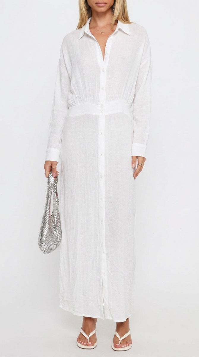 A woman modeling a long white button-front shirt dress with a tie waist, paired with white thong sandals and holding a silver textured handbag against a plain white background.
