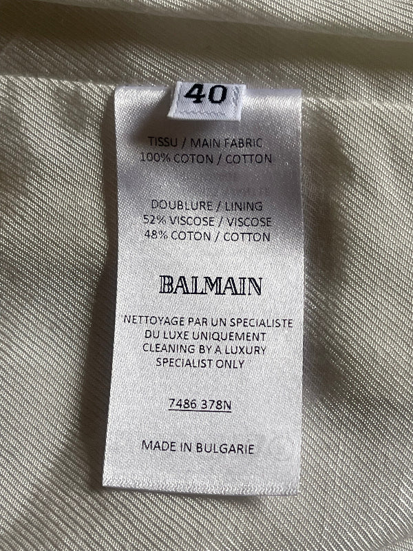 A close-up of a white satin garment tag sewn onto light beige fabric. The tag is branded 'BALMAIN' and shows size '40'.