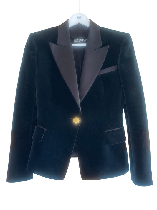 A Balmain black velvet blazer with dark black satin peak lapels and a single gold button, hanging on a white hanger against a white background.