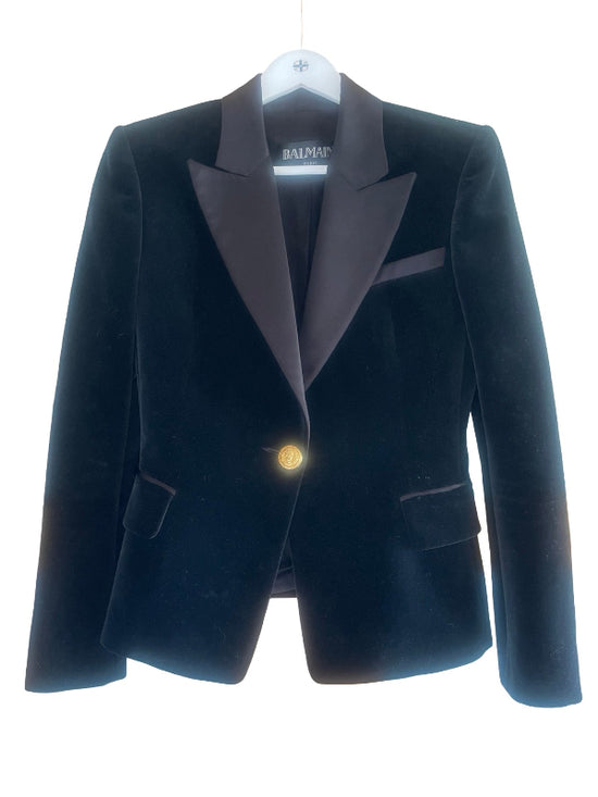 A Balmain black velvet blazer with dark black satin peak lapels and a single gold button, hanging on a white hanger against a white background.