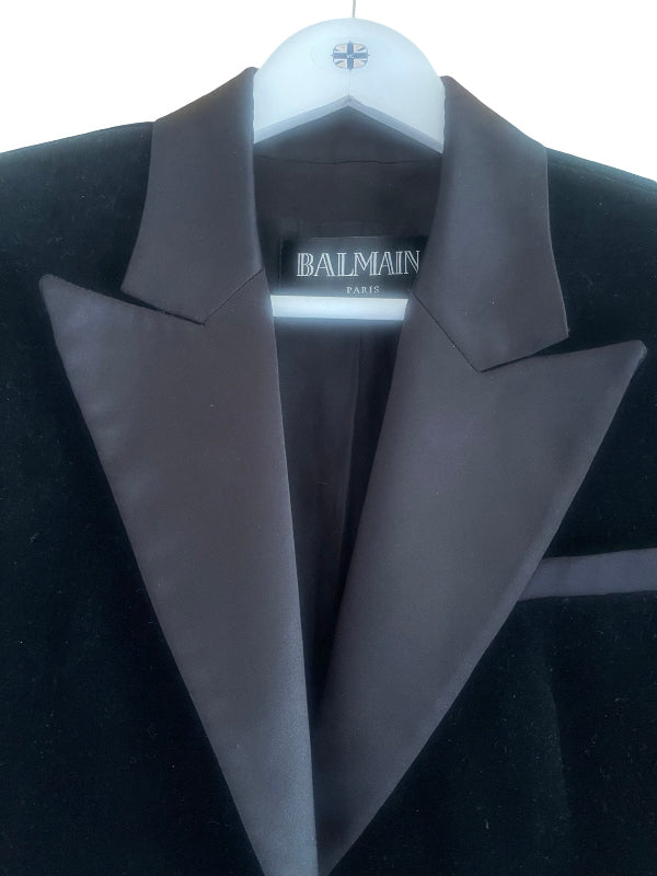 A close-up of a Balmain coat with wide black satin lapels and a black velvet body, hanging on a white hanger against a white background.