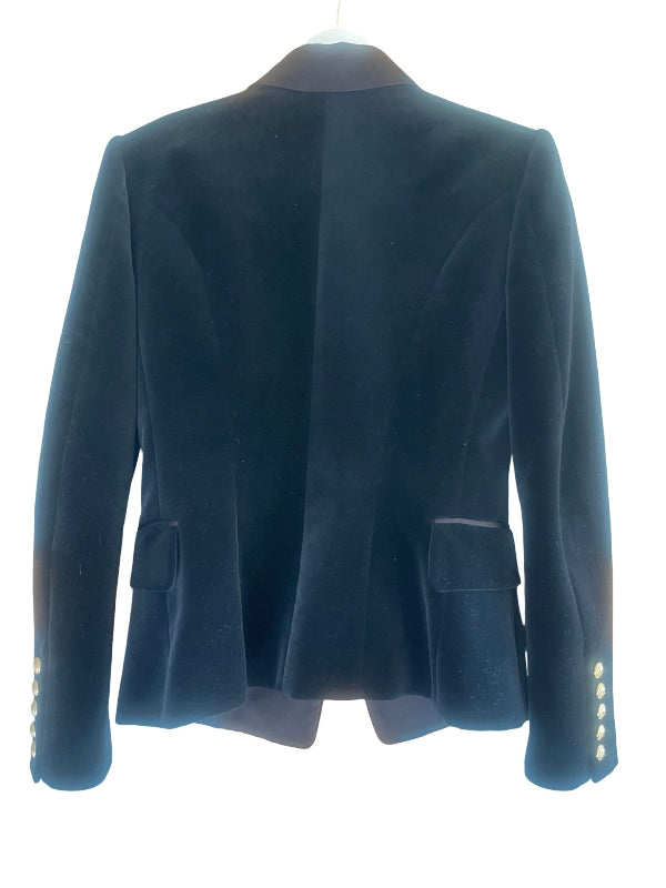 A black velvet blazer shown from the back, featuring structured shoulders, flap pockets, and decorative gold buttons on the cuffs, displayed on a hanger against a white background.