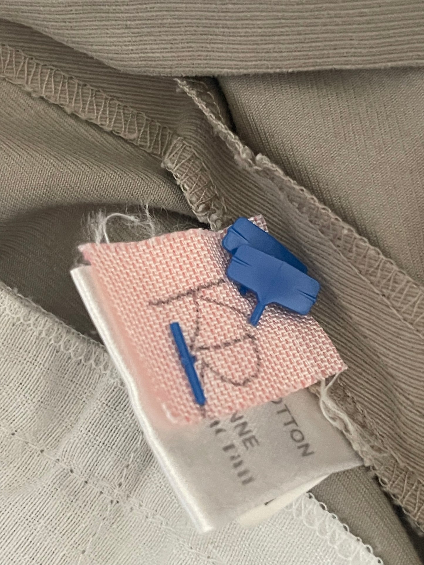 Close-up of a garment's inner label area showing a beige ribbed fabric, a white cotton tag with partial text, and a small pink dry cleaning woven label pinned with blue clips.