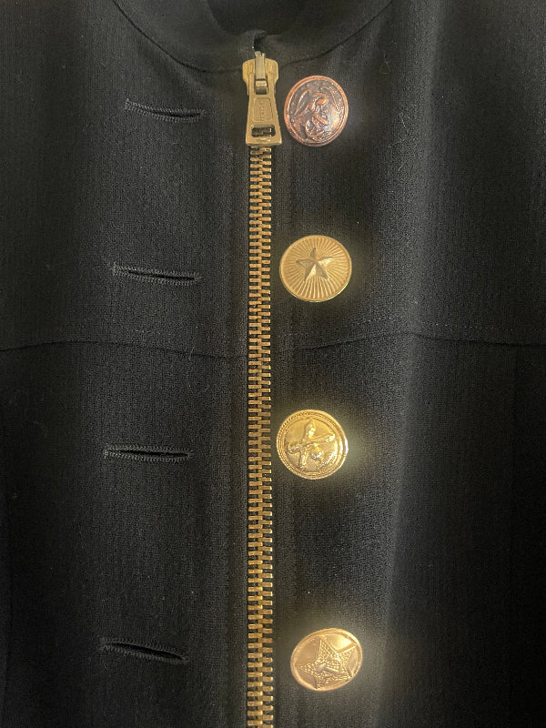 A close-up of a black fabric item featuring a horizontal gold zipper and four matching gold-tone decorative buttons or medallions aligned above it.