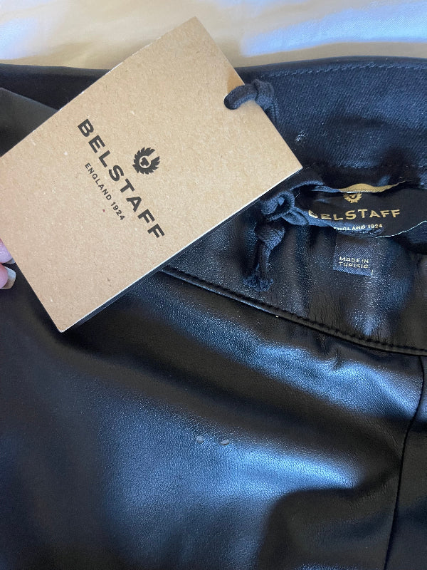 Close-up of black leather or faux-leather fabric, two tiny pin prick flaws with a hanging kraft paper BELSTAFF tag and black drawstring.