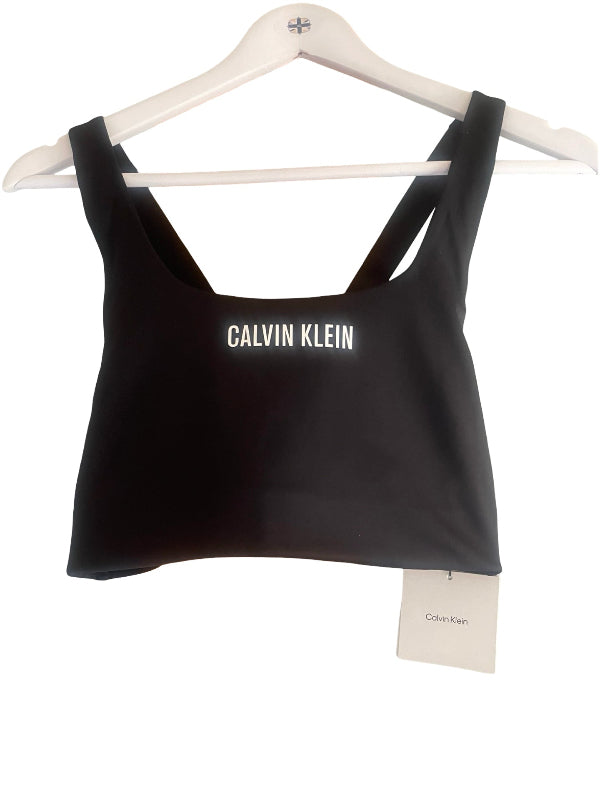 A black Calvin Klein crop top with wide shoulder straps and a white logo printed across the chest, hanging on a white wooden hanger with a Calvin Klein tag attached.