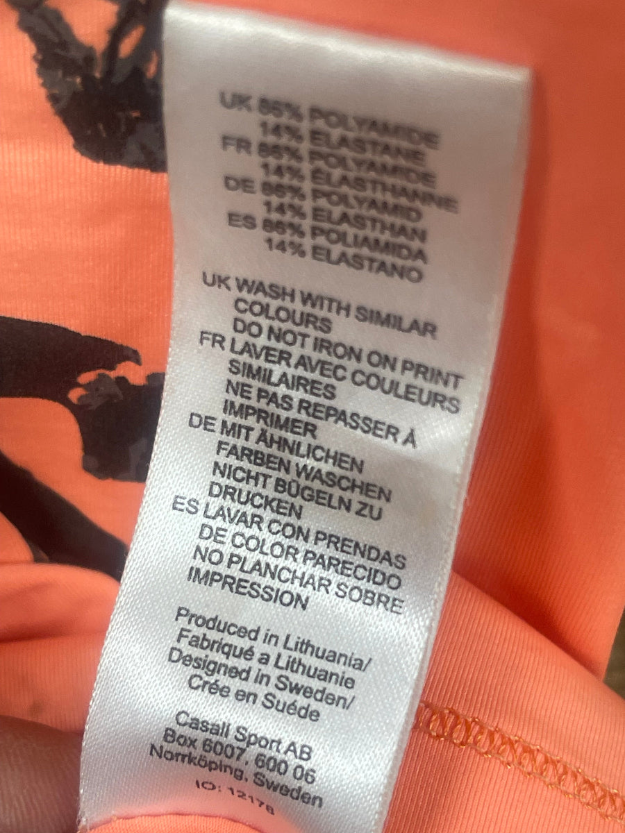 Close-up of a white fabric care label sewn onto an orange garment, showing printed care instructions and country text.