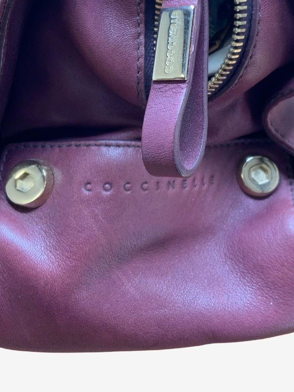 Close-up of a Coccinelle leather bag in a dusty mauve-purple color, showing a gold-tone zipper pull, brass hardware, and the embossed Coccinelle logo on the front.
