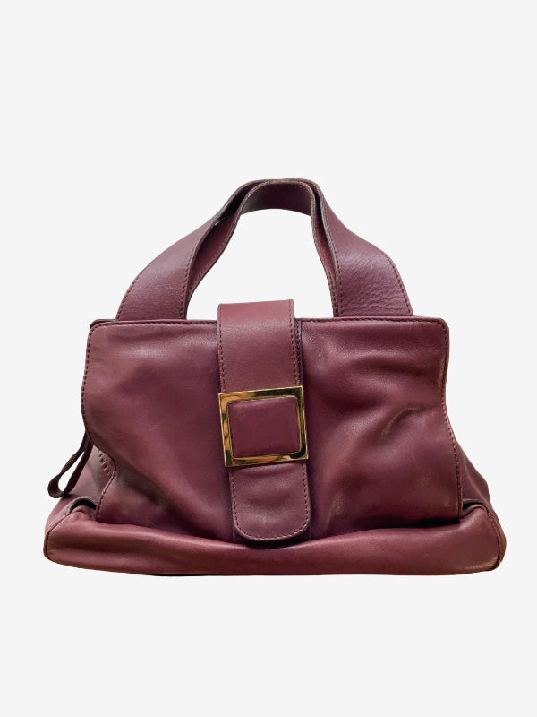 A burgundy leather handbag with a prominent gold-tone buckle closure on the front flap and dual top handles, shown against a plain light gray background.