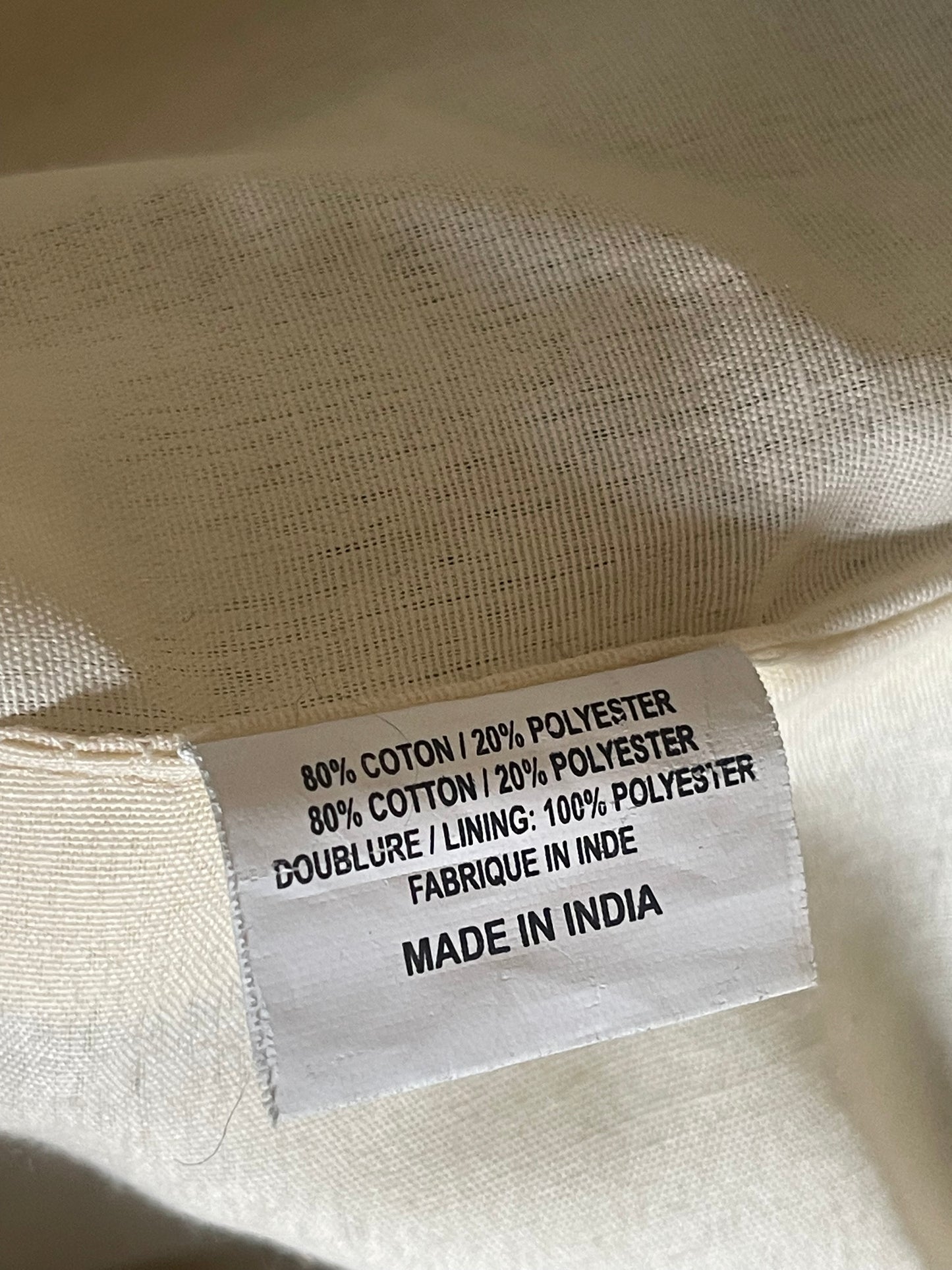 Close-up of a white fabric tag sewn onto the inside collar of a cream-colored garment, showing text that reads '80% COTTON / 20% POLYESTER', '80% COTTON / 20% POLYESTER', '100% POLYESTER', 'DOUBLE LAYER', and 'MADE IN INDIA'.