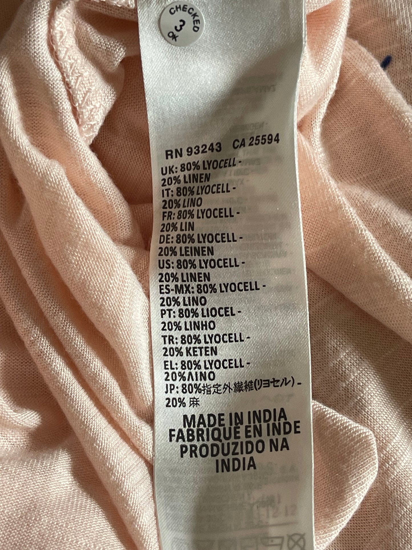 Close-up of a fabric garment label sewn onto light peach-colored textured fabric. The white paper label shows text including '20% Lycell', '20% LIN', '20% LENEN', '20% KETEM', '20% CELL', '20% ALNO', 'FABRICUE EN INDIA', and a circular 'Oversized 3' size sticker.