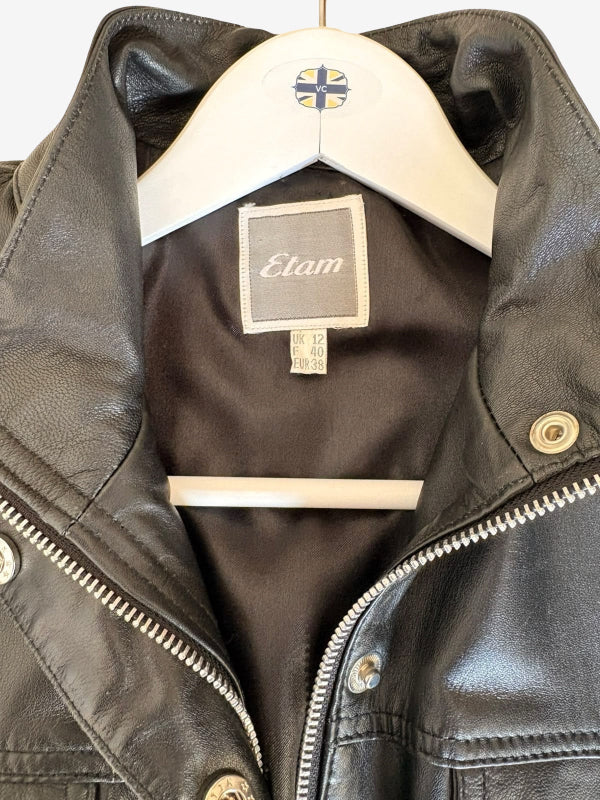 A close-up of a black leather jacket hanging on a white hanger. The jacket features a high collar, silver-toned metal zipper, snap buttons, and a visible interior label reading 'Etam'.