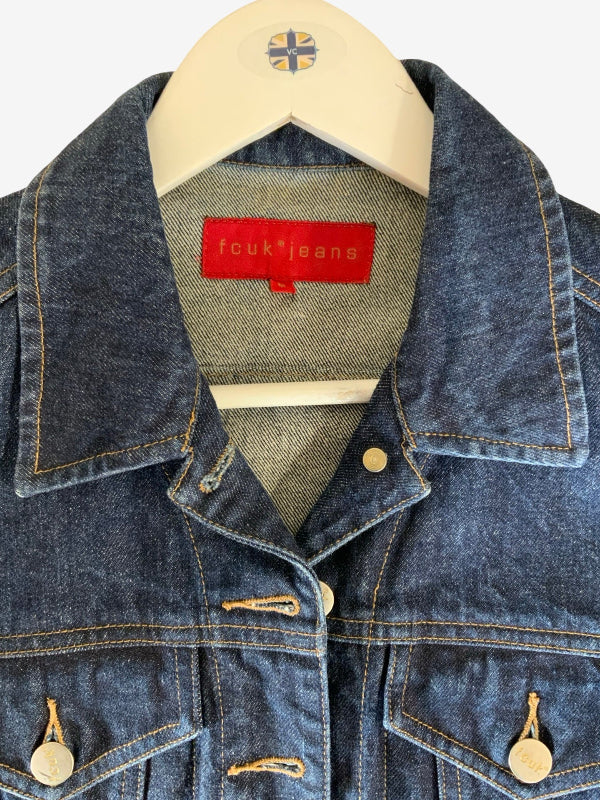 A close-up of a blue denim jacket hanging on a cream hanger. The jacket features a classic collar, buttoned front, and two front flap pockets with brass buttons.