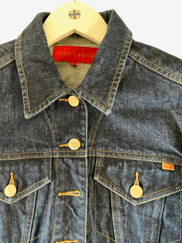 A close-up of a denim jacket hanging on a wooden hanger against a white background. The jacket is made of dark blue denim with yellow contrast stitching, gold-tone metal buttons, and has a red neck label.