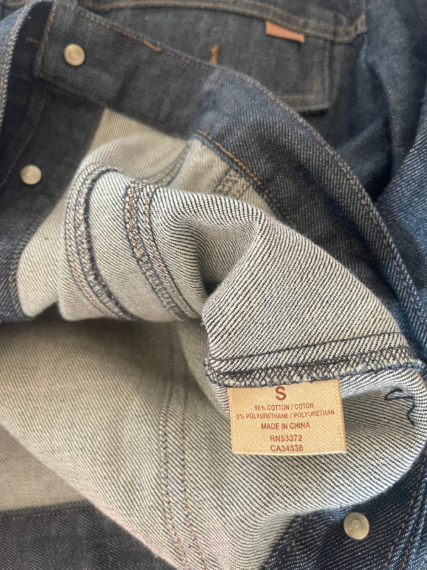 Close-up of folded denim jean jacket showing the inner lining, a sewn-in size label reading S, and metal rivets on a dark blue denim surface.