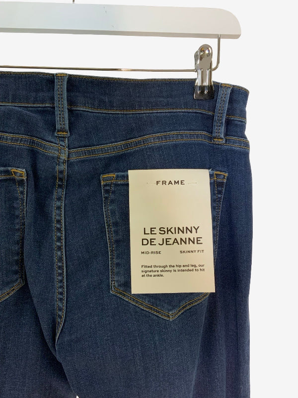 Close-up of the back waistband and pocket area of dark blue denim jeans hanging from a white hanger. A cream paper label on the back pocket reads FRAME and LE SKINNY DE JEANNE.