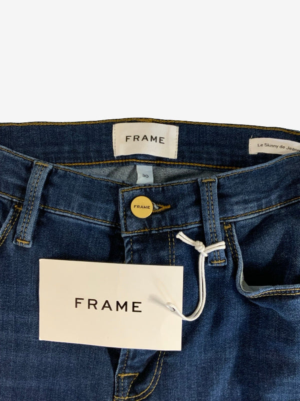 Close-up of the waistband of FRAME denim jeans, showing the blue denim fabric, gold button, white brand labels, and a white paper hangtag.