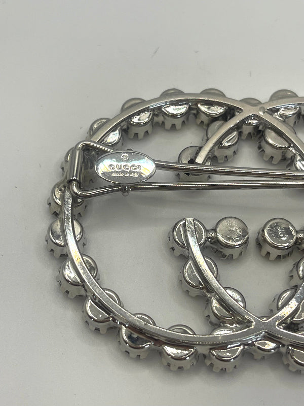 A close-up of a silver-tone Gucci hair clip with the interlocking GG logo, featuring clear rhinestones and a visible Gucci logo stamp on the metal.