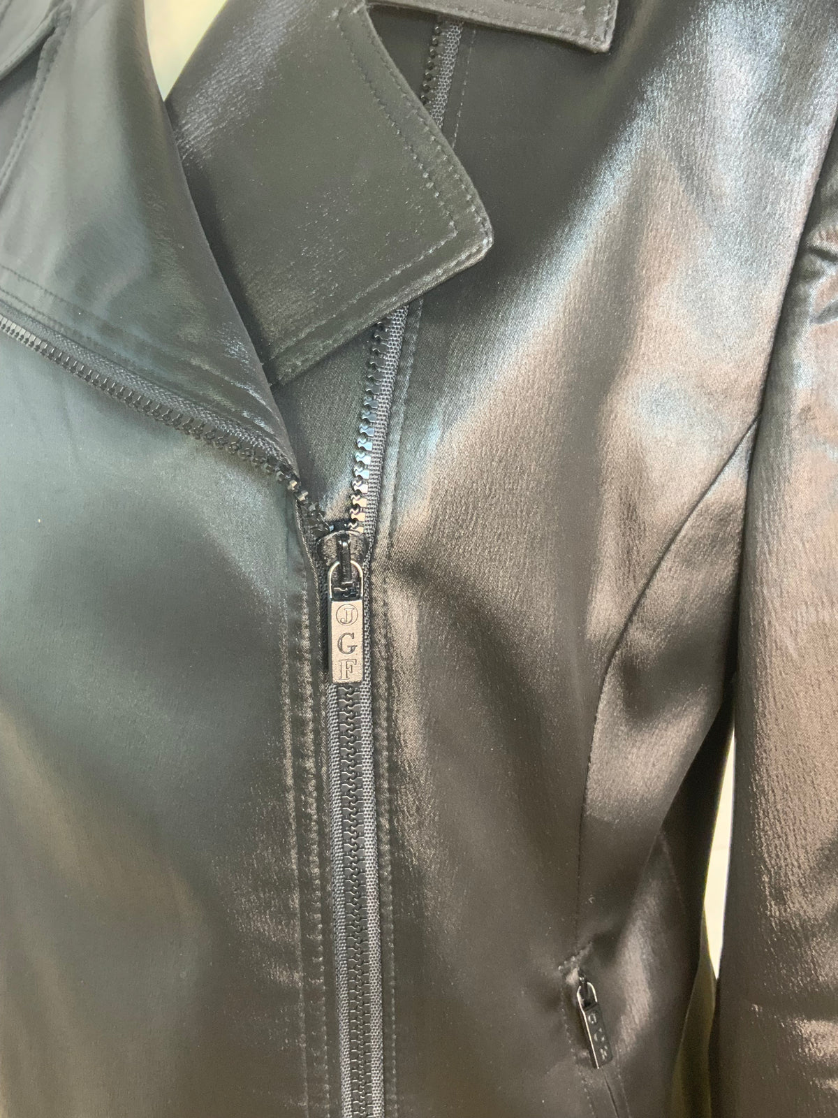 Close-up of a metallic black faux-leather look jacket with a front zipper, collar, and a small metal brand tag reading 'GK'.