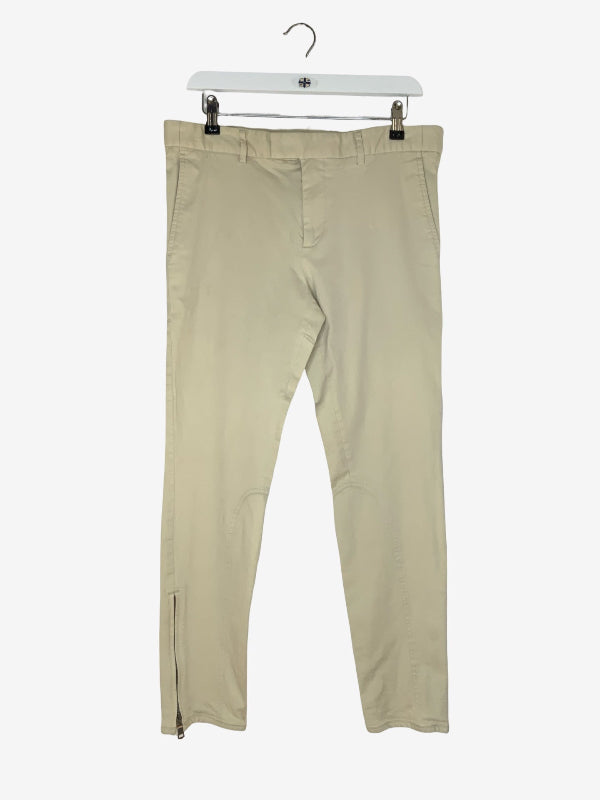 A pair of light beige slim-fit pants with zippered ankle cuffs, hanging from a white plastic hanger against a plain white background.