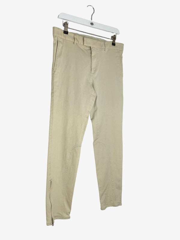 A pair of light beige chino pants hanging from a white plastic hanger with metal clips against a plain white background.