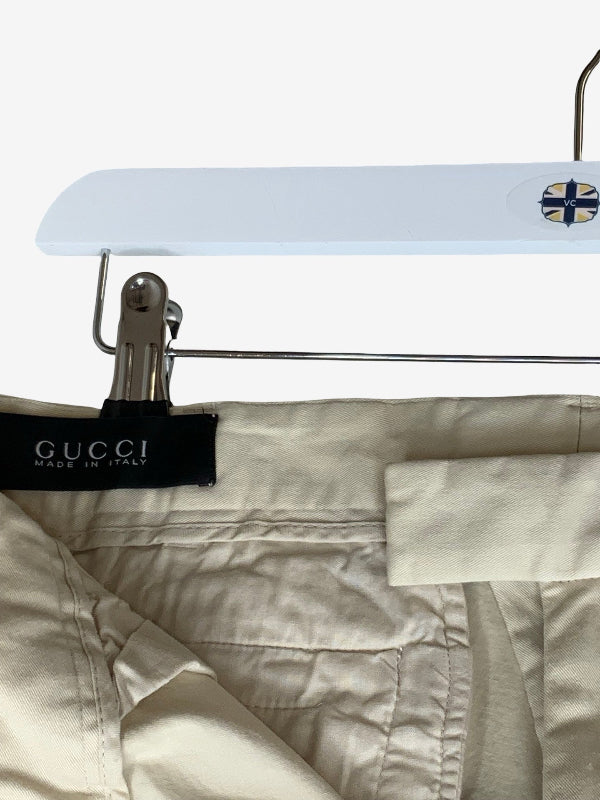 Close-up of Gucci trousers in a light beige or cream color, hanging on a white hanger with a metal clip holding the waistband. The black Gucci brand label is visible on the left side.