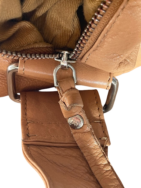 Close-up of a tan leather handbag showing the zipper, metal hardware, and a hanging strap tab with a silver-tone logo stud.