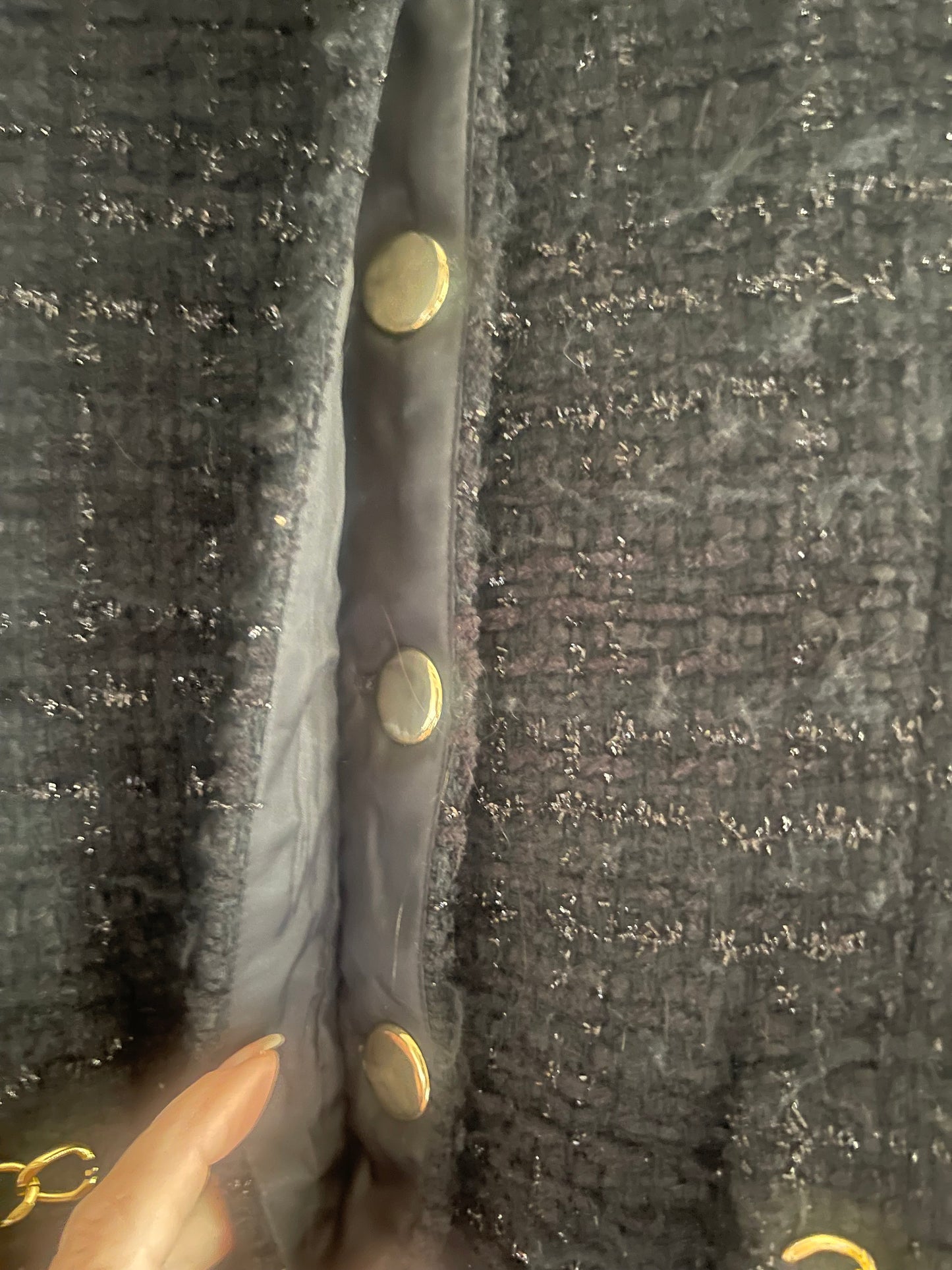Close-up of a dark gray tweed fabric with a subtle shimmer, featuring a horizontal seam with gold-tone snap buttons and a hand pointing to the interior lining.
